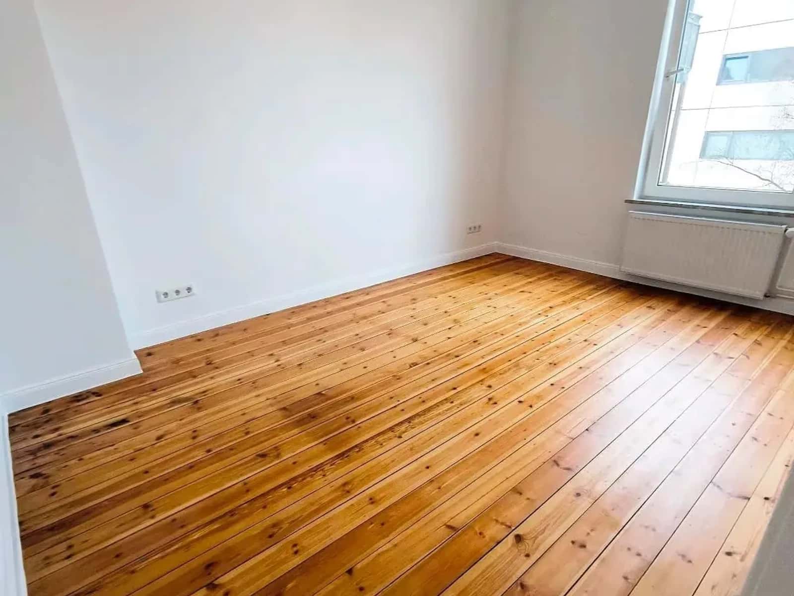 Leeres Altbauzimmer nach der Sanierung: aufgearbeitete Kieferndielen und ein hohes Fenster mit tiefer Laibung