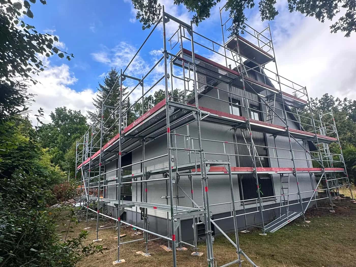 Ein Wohnhaus komplett eingerüstet: Gerüst über drei Ebenen an Fassade und Dach, ringsum Garten und alte Bäume